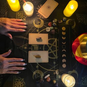 May include: A black cloth with gold and white patterns, featuring a spread of tarot cards, crystals, and candles. The cloth is surrounded by hands, suggesting a spiritual or divination setting.