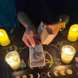 Peut inclure: Une lecture de cartes de tarot avec une bougie allum&eacute;e et une carte du Chevalier de Coupes tir&eacute;e. La carte repr&eacute;sente un chevalier mont&eacute; sur un cheval blanc tenant une coupe dans sa main. La carte est sur une surface noire avec un motif de phases lunaires.