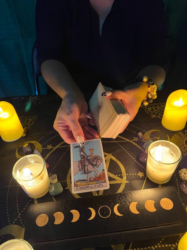 Peut inclure: Une lecture de cartes de tarot avec une bougie allum&eacute;e et une carte du Chevalier de Coupes tir&eacute;e. La carte repr&eacute;sente un chevalier mont&eacute; sur un cheval blanc tenant une coupe dans sa main. La carte est sur une surface noire avec un motif de phases lunaires.