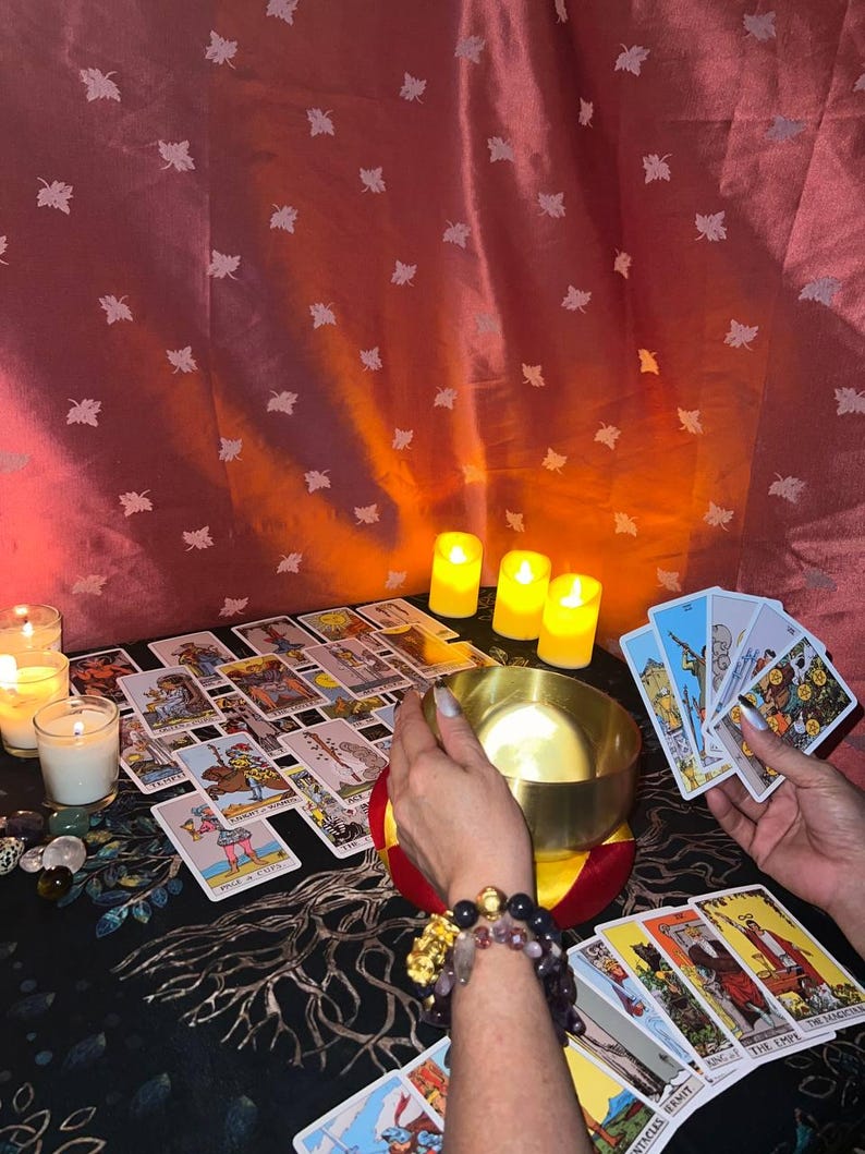 May include: A tarot card reading spread with a variety of cards laid out on a black table cloth with a tree design. The cards are surrounded by candles and a golden bowl. The person is wearing a bracelet with colorful stones.