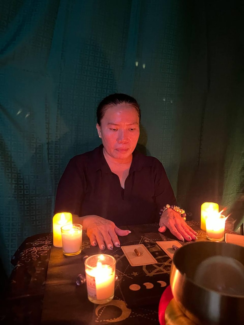 May include: A person sits at a table with a black tablecloth and candles lit. There are tarot cards spread out on the table, along with a bowl and other objects. The person's hands are resting on the table.