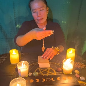 May include: A person is performing a divination ritual with a pendulum, tarot cards, candles, and crystals on a table with a black and gold design. The table is covered with a black cloth with a gold pattern.