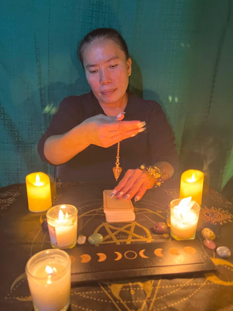 May include: A person is performing a divination ritual with a pendulum, tarot cards, candles, and crystals on a table with a black and gold design. The table is covered with a black cloth with a gold pattern.