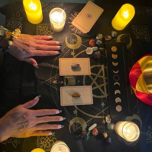 May include: A black cloth with a gold and white pattern, featuring a circle of lit candles, tarot cards, and crystals. The cloth is spread out on a table, and there are two hands in the image.