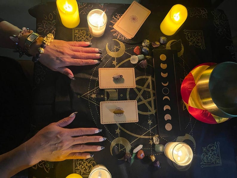 May include: A black cloth with a gold and white pattern, featuring a circle of lit candles, tarot cards, and crystals. The cloth is spread out on a table, and there are two hands in the image.