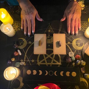 May include: A black cloth with a pentagram design, surrounded by candles, crystals, and two cards with small brown stones on them. The cloth is decorated with moons and stars.
