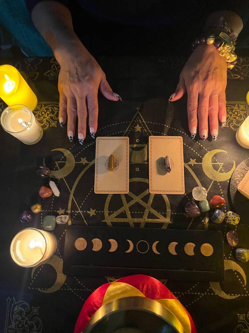 May include: A black cloth with a pentagram design, surrounded by candles, crystals, and two cards with small brown stones on them. The cloth is decorated with moons and stars.