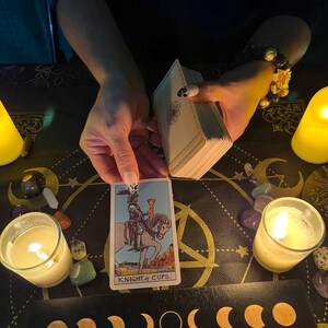 May include: A tarot card reading with a Knight of Cups card pulled. The card is surrounded by candles, crystals, and a moon phase display.