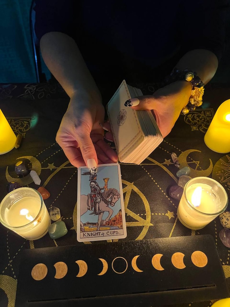 May include: A tarot card reading with a Knight of Cups card pulled. The card is surrounded by candles, crystals, and a moon phase display.