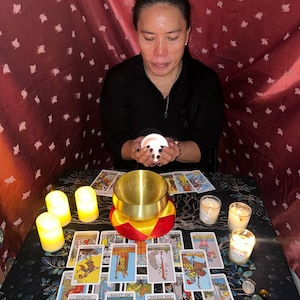 May include: A person sits at a table with a red tablecloth, holding a white candle. The table is covered in tarot cards and candles. There are also several small stones on the table.