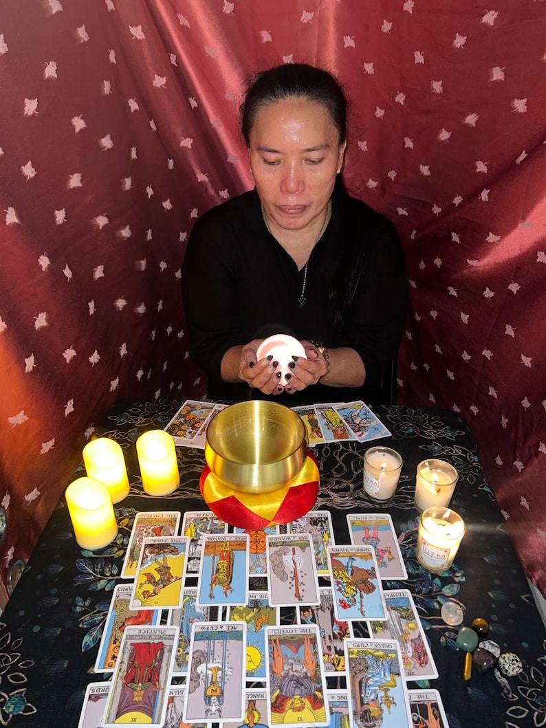 May include: A person sits at a table with a red tablecloth, holding a white candle. The table is covered in tarot cards and candles. There are also several small stones on the table.
