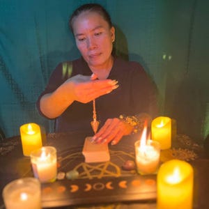 May include: A person sits at a table with candles and crystals, holding a pendulum over a book. The table is covered in a dark cloth with a pentagram design.