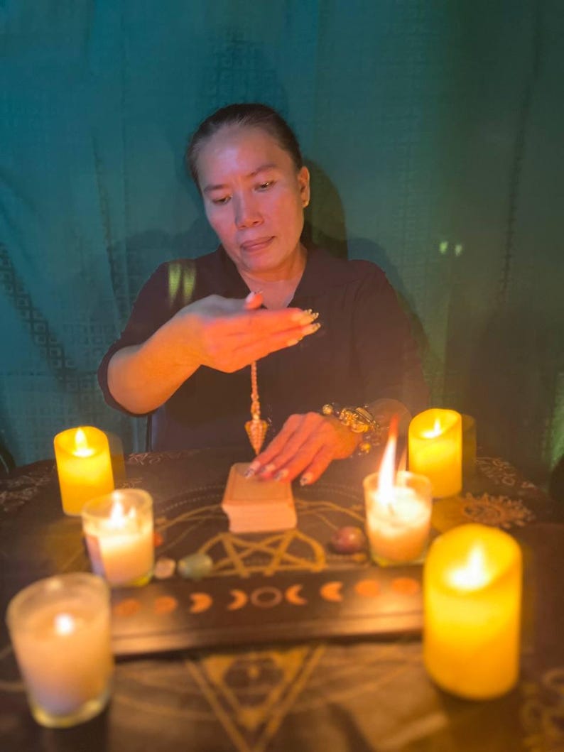 May include: A person sits at a table with candles and crystals, holding a pendulum over a book. The table is covered in a dark cloth with a pentagram design.