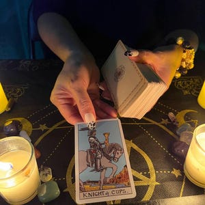 Puede incluir: Una lectura de tarot con la carta del Caballero de Copas tirada. La carta muestra a un caballero montando un caballo blanco, sosteniendo una copa dorada. La carta est&aacute; sobre una superficie negra con un patr&oacute;n dorado y negro.