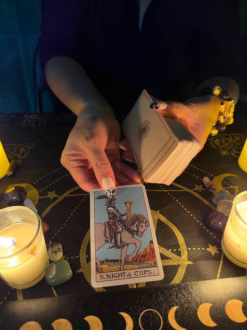 Puede incluir: Una lectura de tarot con la carta del Caballero de Copas tirada. La carta muestra a un caballero montando un caballo blanco, sosteniendo una copa dorada. La carta est&aacute; sobre una superficie negra con un patr&oacute;n dorado y negro.