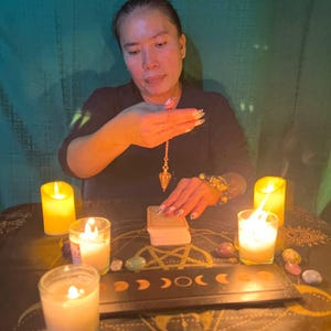 Puede incluir: Una mujer se sienta en una mesa con velas y cristales, sosteniendo un p&eacute;ndulo sobre una baraja de cartas de tarot. La mesa est&aacute; cubierta con una tela oscura con un dise&ntilde;o de pentagrama.