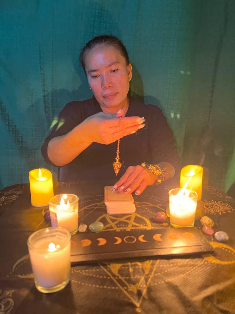 Puede incluir: Una mujer se sienta en una mesa con velas y cristales, sosteniendo un p&eacute;ndulo sobre una baraja de cartas de tarot. La mesa est&aacute; cubierta con una tela oscura con un dise&ntilde;o de pentagrama.