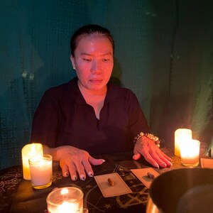 May include: A woman sits at a table with three lit candles, a bowl, and tarot cards spread out. The table has a black and gold patterned tablecloth.