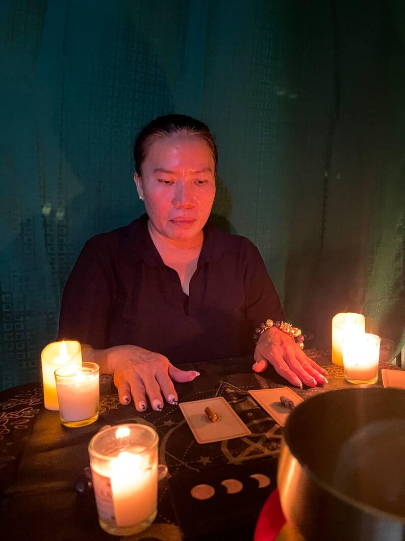 May include: A woman sits at a table with three lit candles, a bowl, and tarot cards spread out. The table has a black and gold patterned tablecloth.