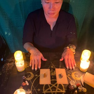 May include: A person sits at a table with two lit candles, crystals, and two tarot cards. The cards are on a black surface with a gold design of stars, moons, and a pentagram. The cards have a gold border and a white background.