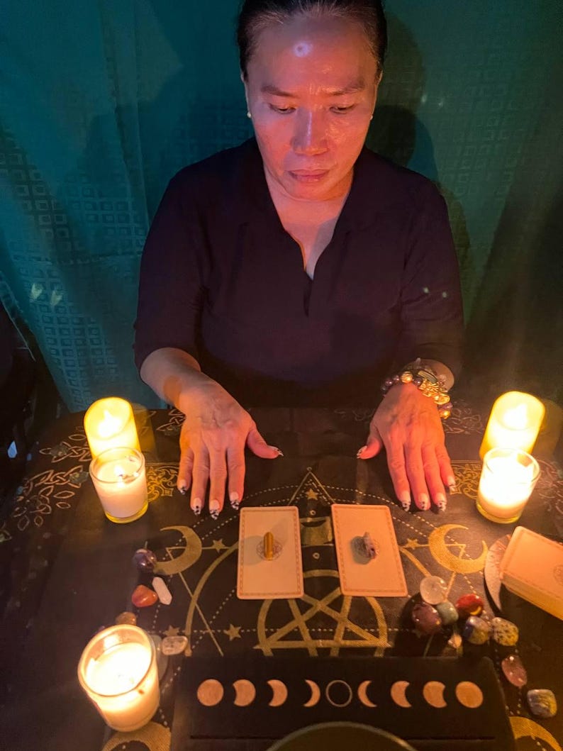 May include: A person sits at a table with two lit candles, crystals, and two tarot cards. The cards are on a black surface with a gold design of stars, moons, and a pentagram. The cards have a gold border and a white background.