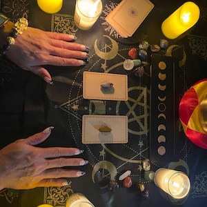 May include: A black altar cloth featuring gold celestial patterns, two rectangular tarot cards with crystals placed on top, a moon phase chart, scattered polished gemstones, three lit pillar candles, and a gold singing bowl with a red and yellow cushion.