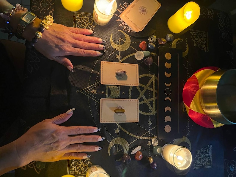 May include: A black altar cloth featuring gold celestial patterns, two rectangular tarot cards with crystals placed on top, a moon phase chart, scattered polished gemstones, three lit pillar candles, and a gold singing bowl with a red and yellow cushion.