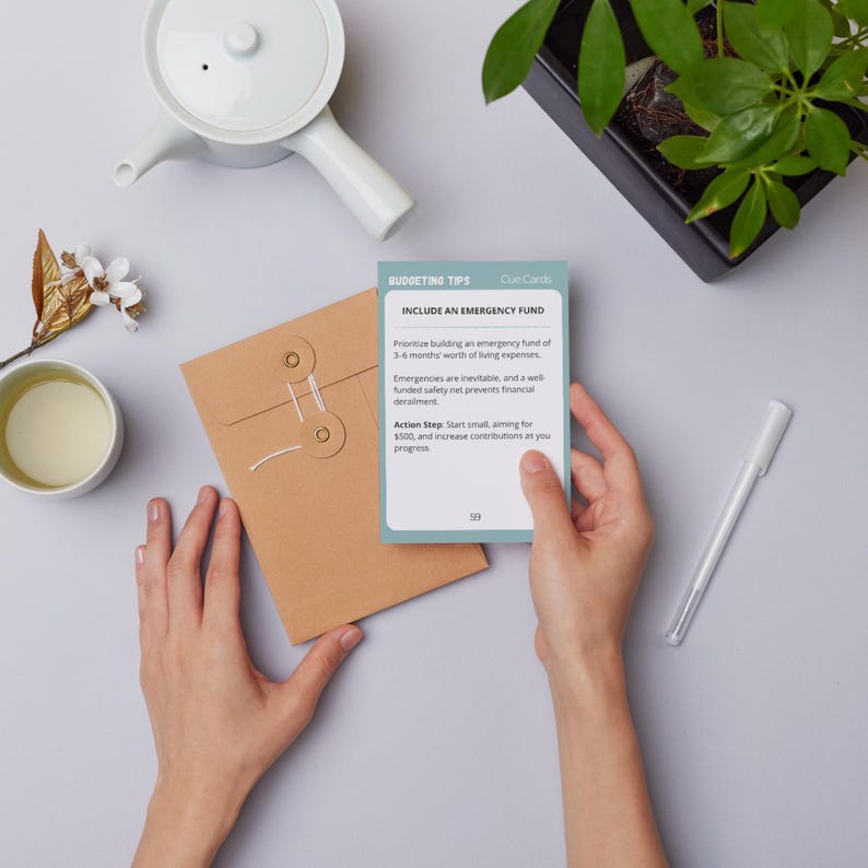 May include: A light blue and green card with the text "BUDGETING TIPS" and "INCLUDE AN EMERGENCY FUND". The card has a white border and the text is in black. The card is being held in a person's hand.