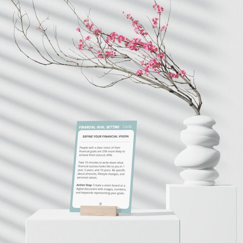 May include: A blue and white card with the text "Financial Goal Setting Cards" and "Define Your Financial Vision". The card has a light blue background and white text. The card is sitting on a wooden stand.