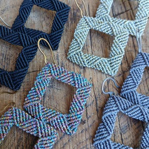 Peut inclure: Gros plan sur des boucles d'oreilles macramé faites à la main, de couleurs variées : noir, argent, irisé et gris. Chaque boucle d'oreille présente un motif carré avec une texture tissée et un crochet métallique. Présentées sur une surface en bois.