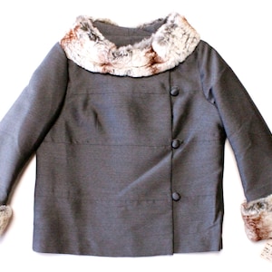 Puede incluir: Una chaqueta gris con cuello y puños de piel sintética. La chaqueta tiene cierre de botón y un diseño ligeramente asimétrico.