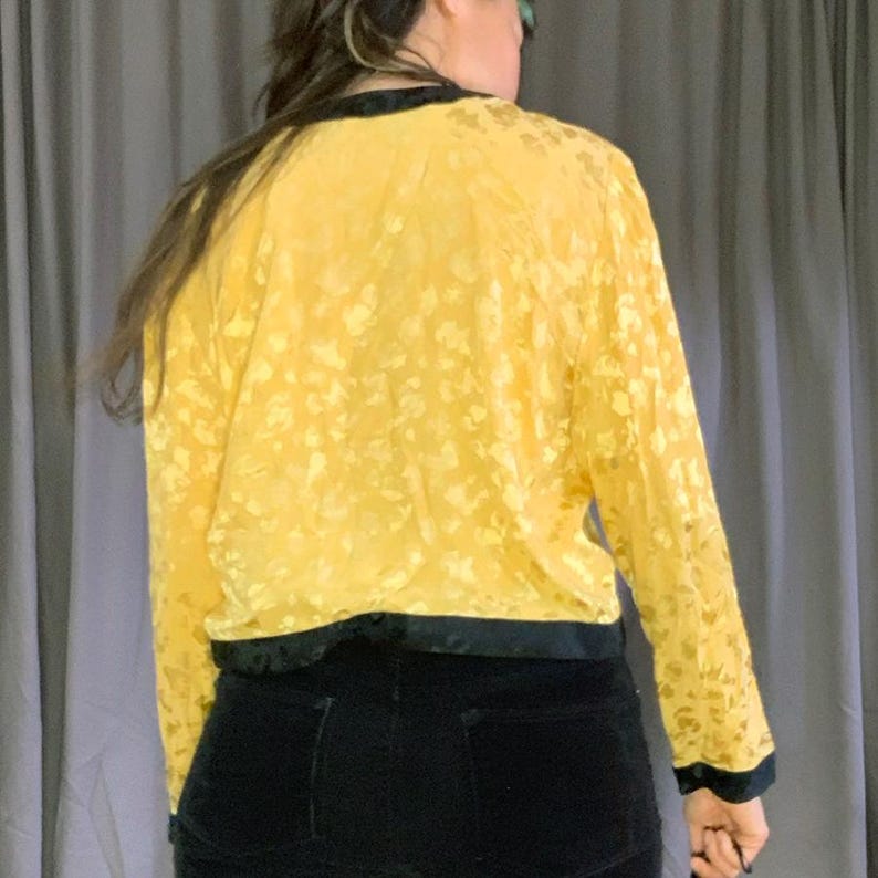 May include: A yellow floral-patterned jacket with black trim. The long-sleeved jacket has a satin-like texture and is paired with black trousers. The back view of the jacket shows the floral pattern and the black trim around the collar and cuffs.