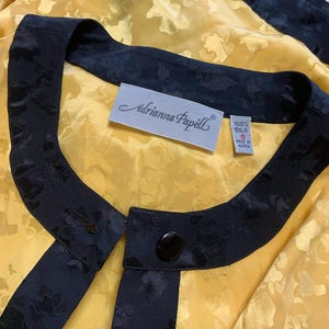 May include: A vintage yellow silk blouse with a floral pattern, featuring black trim and a button closure. The label reads "Adrianna Papell" and indicates the blouse is size 6 and made of 100% silk.