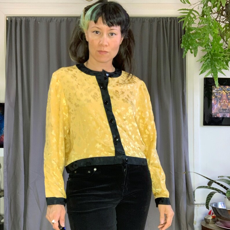 May include: A yellow blouse with a floral pattern and black trim. The long-sleeved top has a round neckline and is buttoned down the front. The trim is around the collar, cuffs, and bottom hem. The blouse is paired with black velvet trousers.