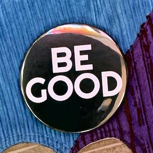 Può includere: Un bottone rotondo nero con le parole "BE GOOD" in grandi lettere bianche. Il bottone ha una superficie lucida e riflettente, su uno sfondo blu e viola.