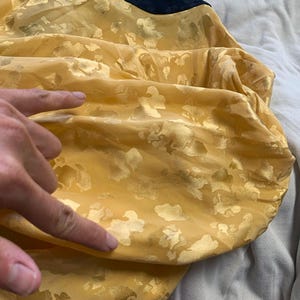 May include: Close-up of a yellow fabric with a gold floral pattern. The fabric is gathered, creating folds and shadows. The texture appears soft and slightly translucent. A dark blue fabric is visible at the top.