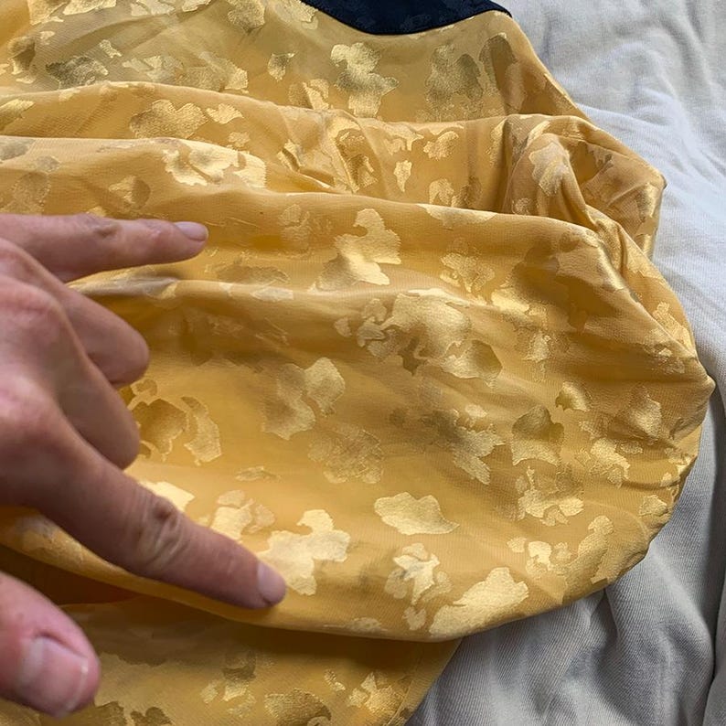 May include: Close-up of a yellow fabric with a gold floral pattern. The fabric is gathered, creating folds and shadows. The texture appears soft and slightly translucent. A dark blue fabric is visible at the top.
