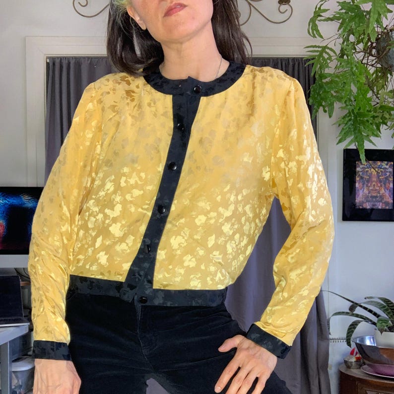 May include: A vintage yellow blouse with a floral pattern and black trim. The long-sleeved top has a round neckline and a button-up front. The black trim is on the collar, down the front, and at the cuffs and hem. The blouse is made of a shiny material.
