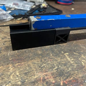 Puede incluir: Primer plano de un soporte de reparaci&oacute;n de bicicletas de metal negro y azul. El soporte tiene una base negra con un dise&ntilde;o en forma de cruz y un brazo curvo azul. El soporte est&aacute; sobre una superficie de madera.