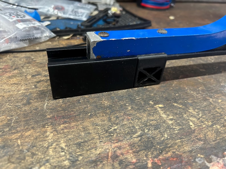 Puede incluir: Primer plano de un soporte de reparaci&oacute;n de bicicletas de metal negro y azul. El soporte tiene una base negra con un dise&ntilde;o en forma de cruz y un brazo curvo azul. El soporte est&aacute; sobre una superficie de madera.