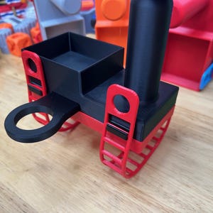 May include: A 3D printed toy train engine featuring a black rectangular body with a cylindrical smokestack and a circular hitch attachment. The base is red with two red ladder-like track components on the sides, accented by small black rectangular connectors.