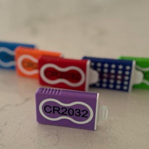 May include: A purple plastic battery holder with a white infinity-shaped outline and the text CR2032 printed in black. Several other battery holders in blue, orange, red, and green are arranged in the background, each featuring the same design.