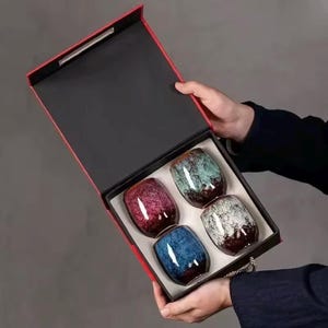 May include: A set of four ceramic tea cups in a red and black gift box. The cups are oval-shaped with a speckled glaze in shades of red, blue, green, and white. The box is held open, showcasing the cups.