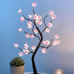 May include: An LED cherry blossom tree with a black base and branches, adorned with pink, illuminated flowers. A small succulent in a grey pot and a lit candle in a glass jar are also on the table.