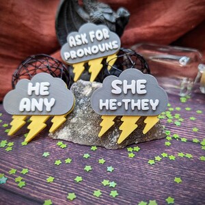 Può includere: Tre spille a forma di nuvola grigia con fulmini gialli. Le spille dicono "Chiedi i pronomi", "Lui Qualsiasi", e "Lei Lui-Loro".