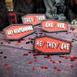 May include: Four red coffin-shaped pins with black trim. Each pin has white text on it. The pins read "They/Them", "She/Her", "Any Neopronouns", and "He/They/She".