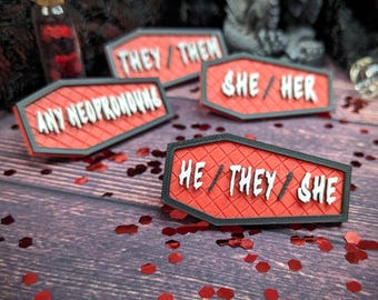 May include: Four red coffin-shaped pins with black trim. Each pin has white text on it. The pins read "They/Them", "She/Her", "Any Neopronouns", and "He/They/She".