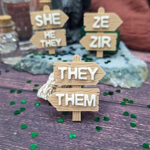 May include: Wooden signposts with the words "She, He, They", "Ze, Zir", and "They, Them" written on them. The signposts are brown and have white lettering.