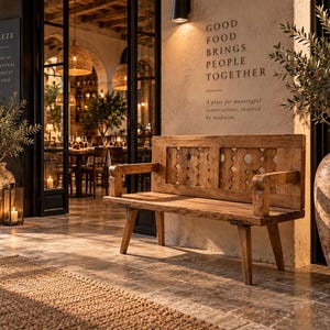 May include: A wooden bench with a rectangular backrest featuring a geometric cutout pattern, positioned on a stone patio. The wall behind the bench displays the text: GOOD FOOD BRINGS PEOPLE TOGETHER. A place for meaningful conversations, inspired by tradition.