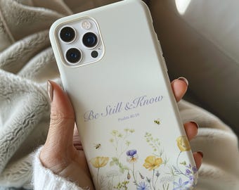 May include: A light-colored phone case with the text "Be Still & Know" and Psalm 46:10. The case features a floral design with yellow, purple, and blue flowers, along with small bees.